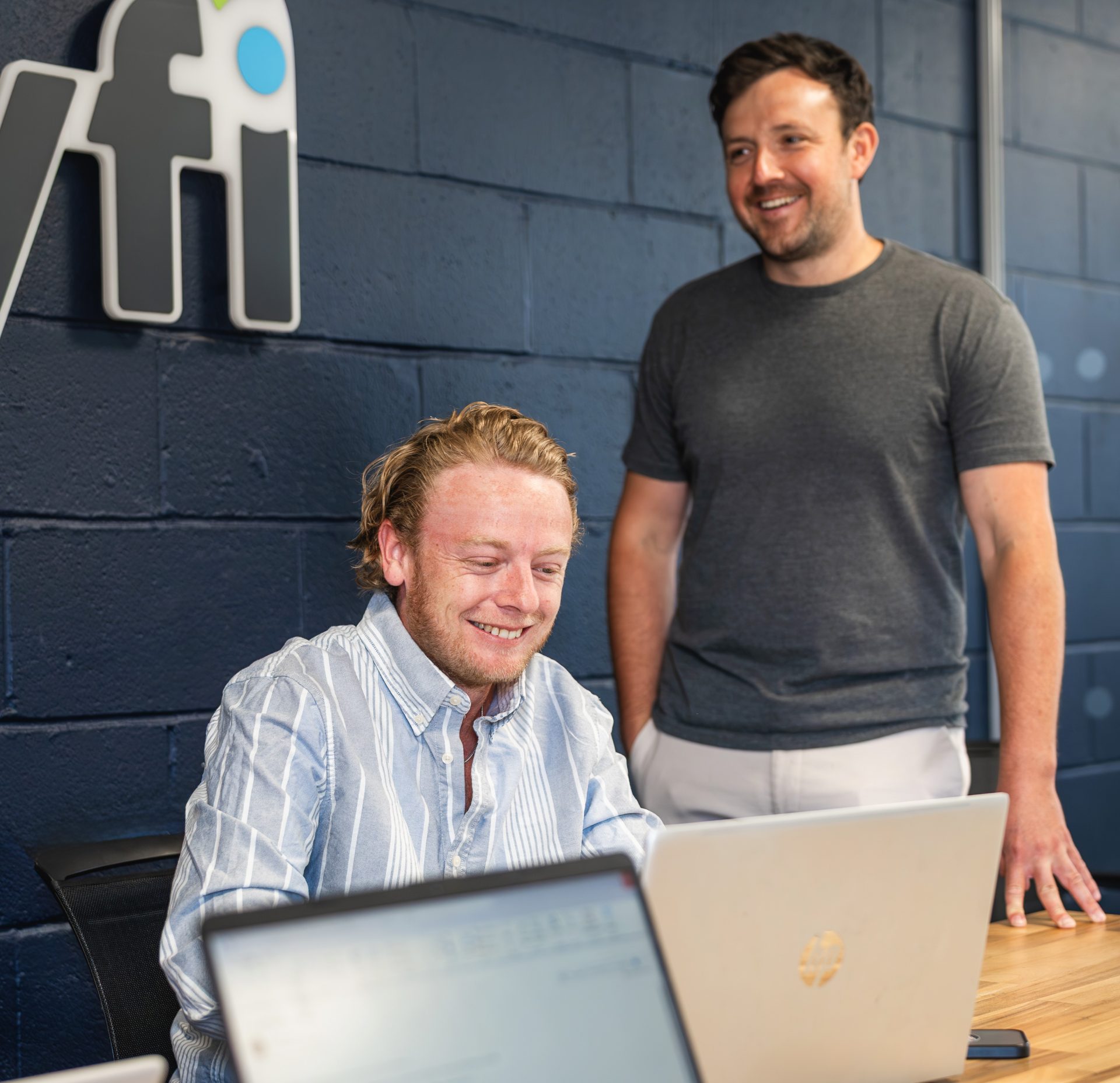 One man sitting down and one man standing and laughing while working on laptops